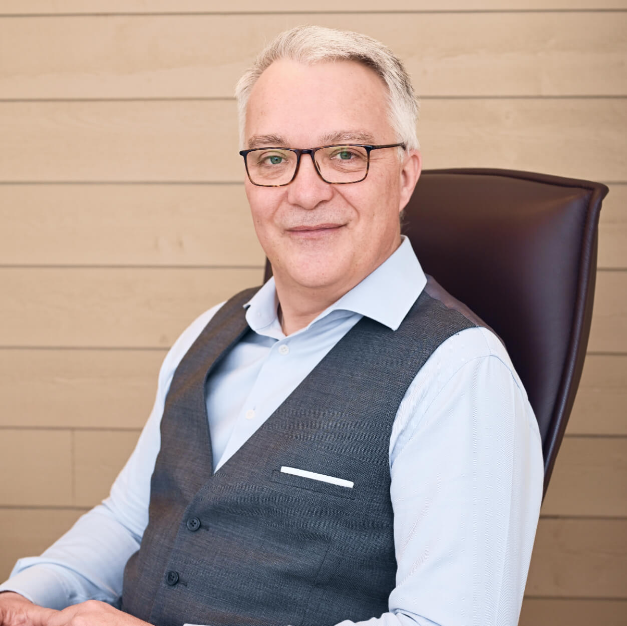 Portrait professionnel de Lionel GUÉRIN, directeur du cabinet de recrutement Learn2Fly RH, en gilet et chemise bleue, assis dans un fauteuil de bureau en cuir marron.
